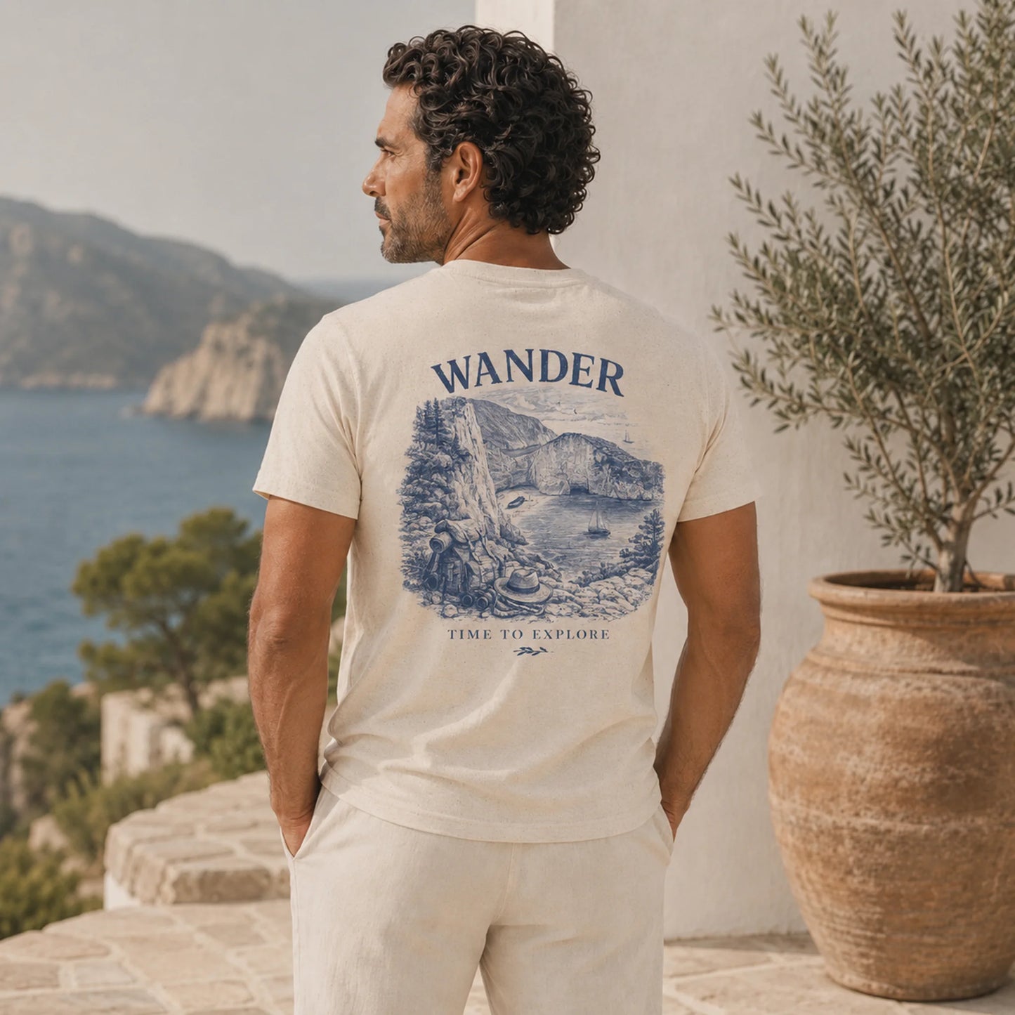 Man wearing a t-shirt with 'Wander' and mountain graphic, standing by a scenic coastal view.