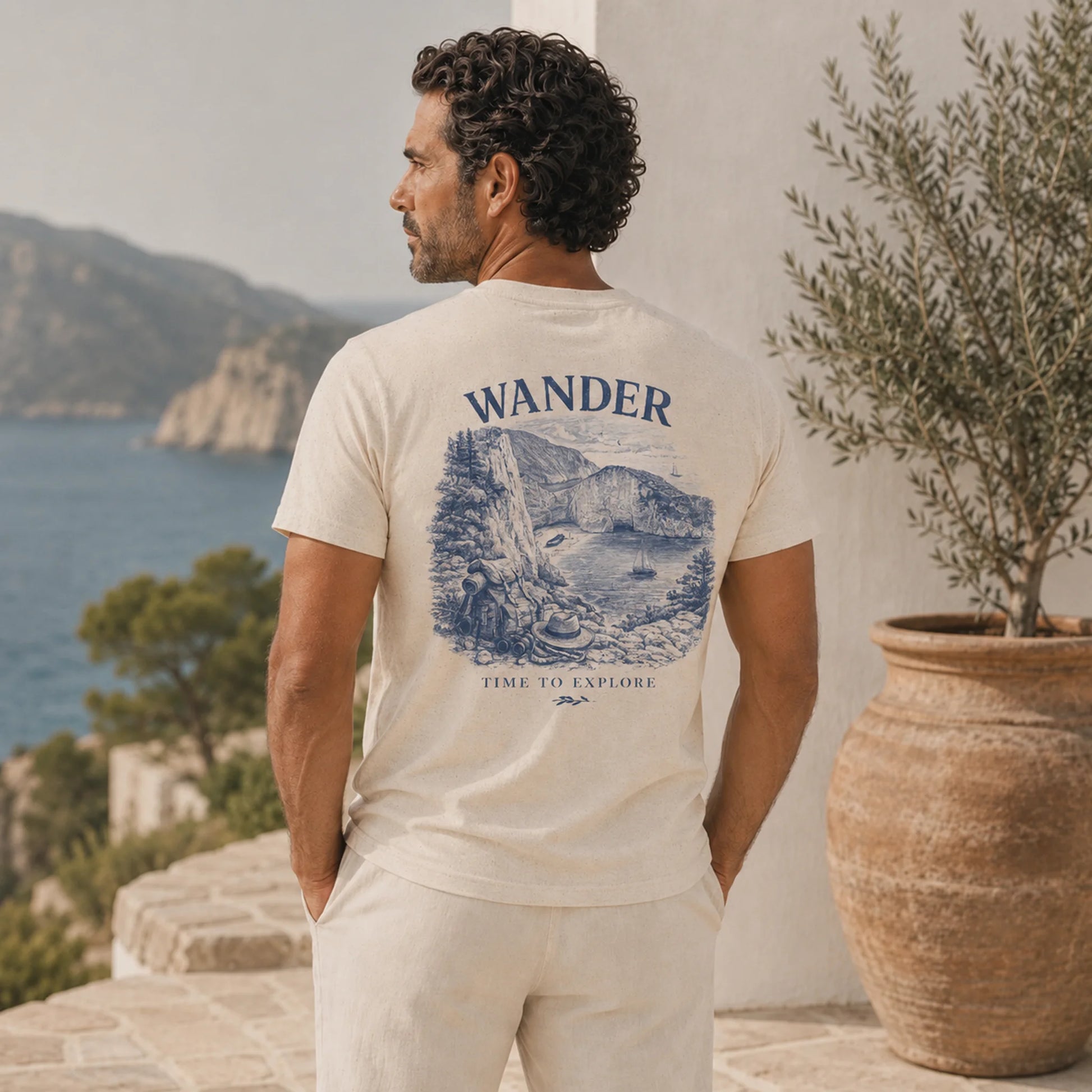 Man wearing a t-shirt with 'Wander' and mountain graphic, standing by a scenic coastal view.