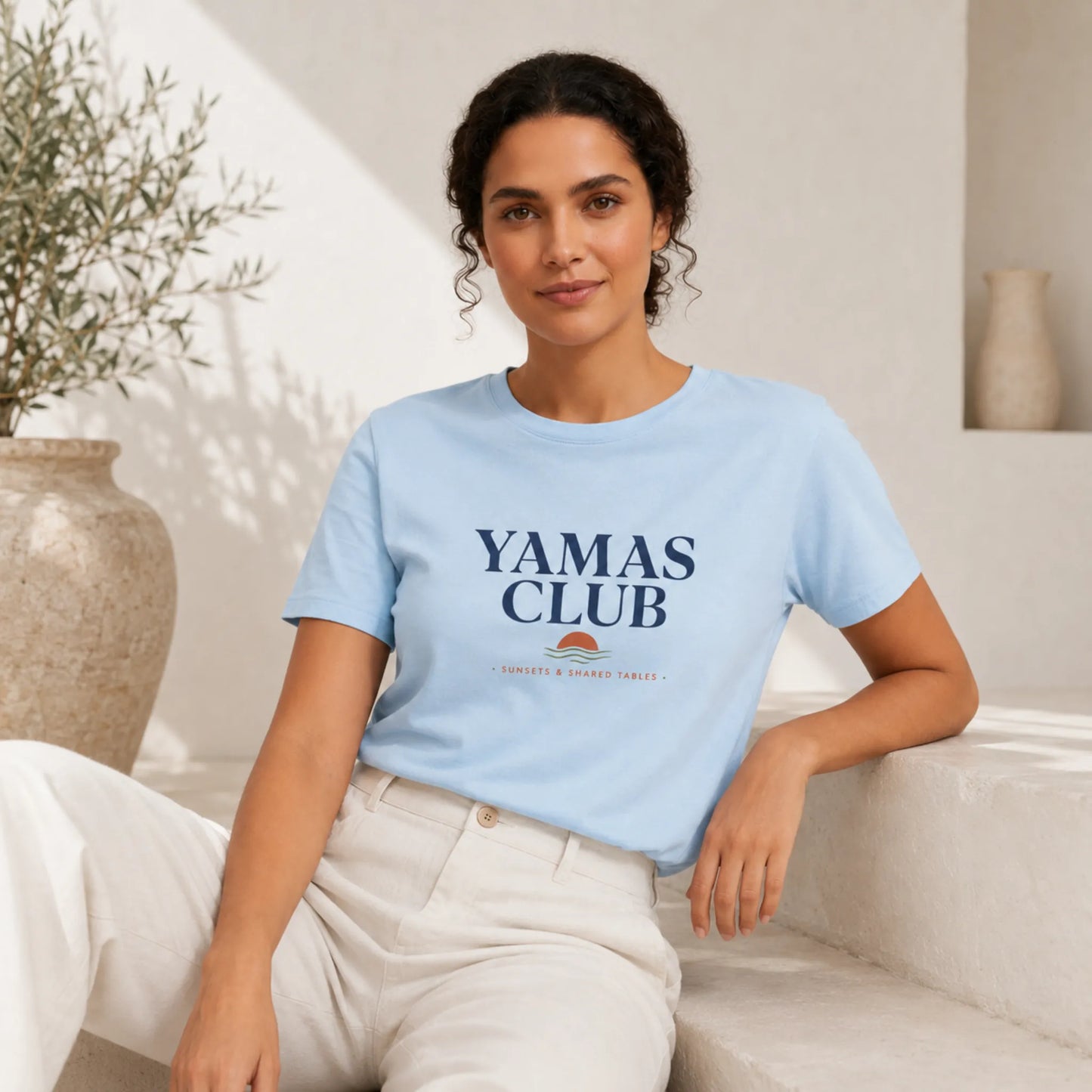 Woman wearing a light blue t-shirt with 'Yamas Club' text, sitting on a couch.